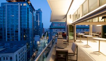 A modern balcony with tables and chairs overlooks a city street lined with tall buildings in the evening light.