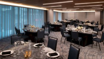 A modern banquet hall with round tables set for an event, featuring elegant place settings, candles, and floral arrangements, lit by overhead lighting.