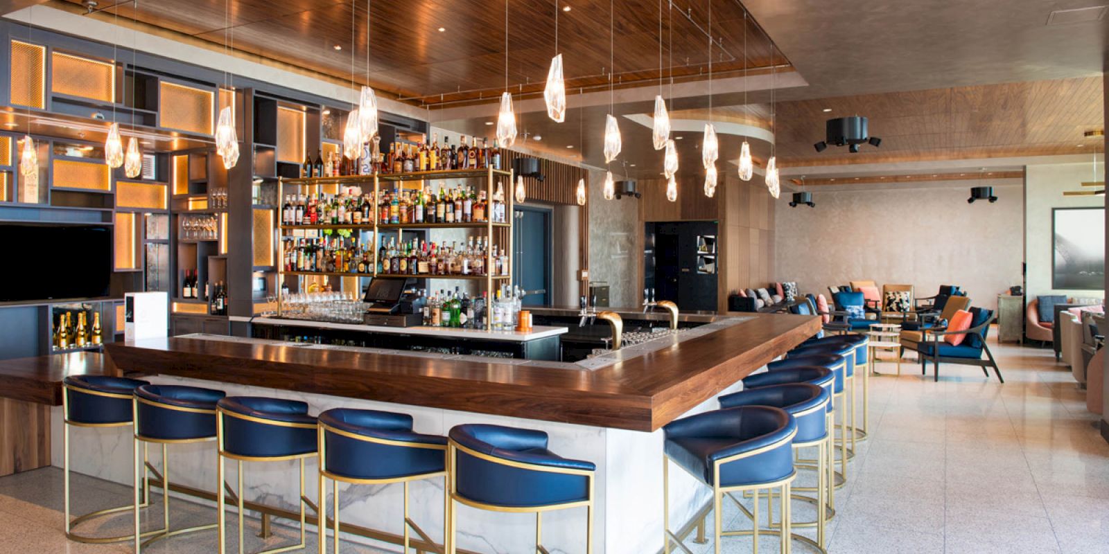 A modern bar with a wooden counter, blue chairs, hanging lights, and a well-stocked back bar, set in a spacious lounge area with additional seating.