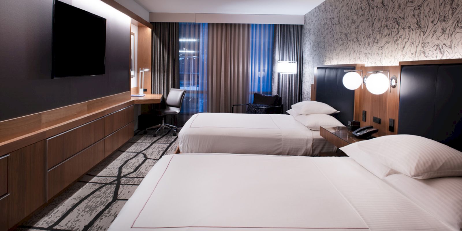 A hotel room with two beds, a flat-screen TV, a desk with a chair, bedside tables, and modern decor is shown in the image.