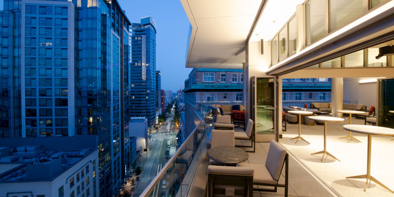 The image shows a modern balcony with seating and tables overlooking a cityscape with tall buildings at dusk, and a street below.