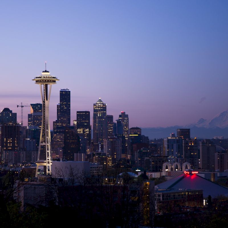 A city skyline at dusk featuring the Space Needle and several skyscrapers, with a snow-capped mountain in the background and a clear night sky.