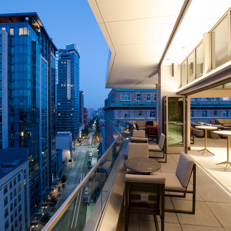A modern balcony with seating, tables, and city skyscraper views during dusk. The indoor area is well-lit with sleek, contemporary furniture.