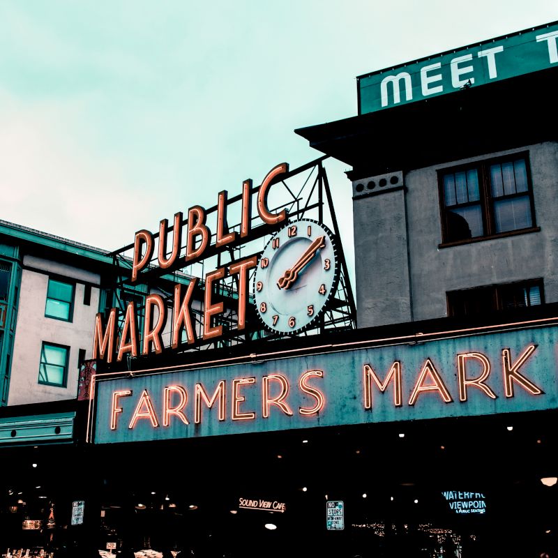 A large neon sign for a public market and farmers market, with a clock in the middle, is displayed on a building in an urban setting.