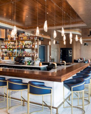 A modern bar with blue and gold bar stools, a variety of bottles on shelves behind the counter, and hanging light fixtures.