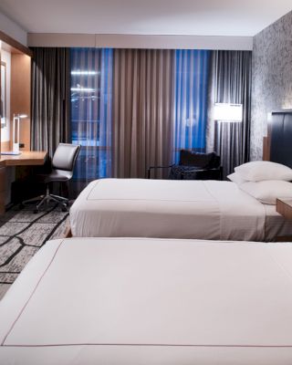 A modern hotel room with two beds, wall-mounted TV, desk, chair, lamps, and intricate wallpaper is shown.