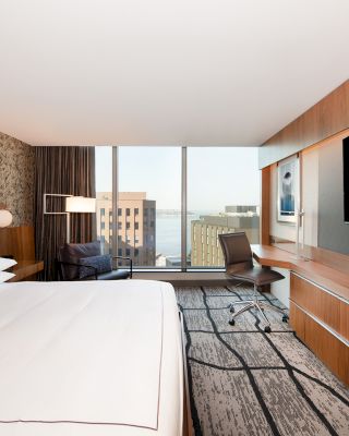 A modern hotel room features a large bed, desk, chair, flat-screen TV, and a window with a cityscape view.