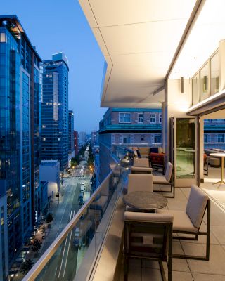 A modern cityscape at dusk with high-rise buildings, viewed from a spacious terrace with tables and chairs. The scene exudes urban elegance.