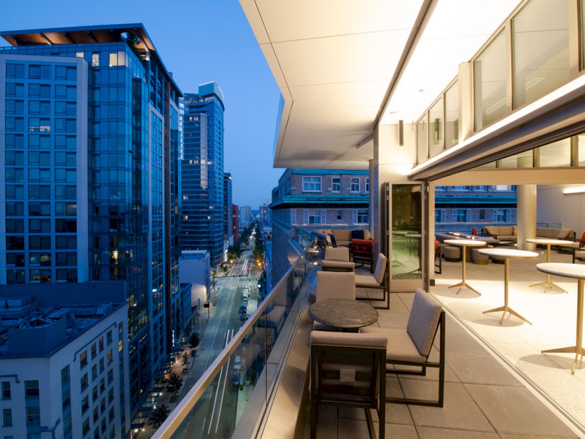 A modern cityscape at dusk with tall buildings, viewed from a spacious terrace with seating and tables, featuring a blend of natural and artificial lighting.