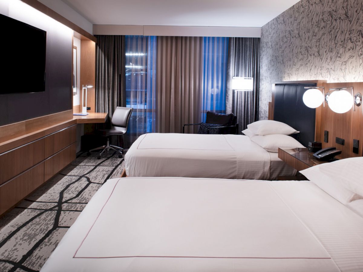 A modern hotel room features two double beds, a flat-screen TV, a desk with a chair, nightstands with lamps, and large windows with curtains.