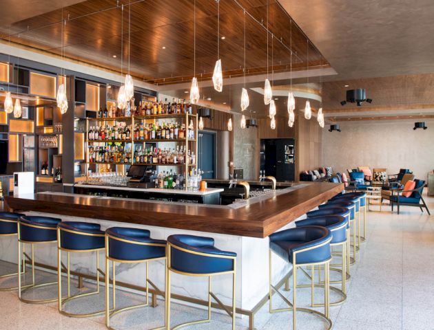 A modern bar with a large selection of drinks, stylish lighting fixtures, blue barstools with gold accents, and a relaxed seating area in the background.