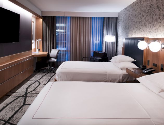A modern hotel room with two beds, a wall-mounted TV, a desk with a chair, and a patterned carpet. The room is well-lit and features contemporary decor.