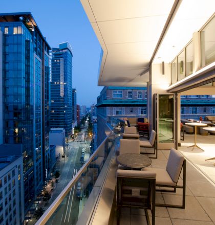 A modern cityscape at dusk with high-rise buildings and a spacious balcony featuring seating and tables overlooking a vibrant urban street.