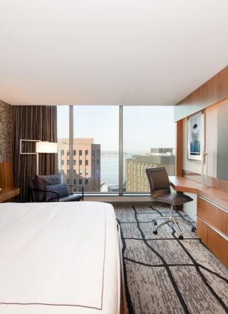 A modern hotel room with a king-sized bed, large TV, desk, and a view of nearby buildings and water through large windows.