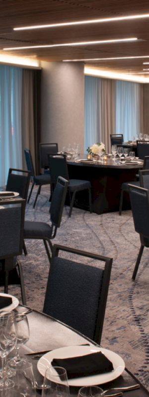 A large, elegantly set dining room with round tables, black chairs, white floral centerpieces, and candles, with dim overhead lighting and blue curtains.