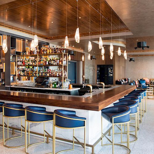 A modern bar with a sleek design, featuring a wooden countertop, blue chairs, hanging lights, and shelves stocked with various drinks.