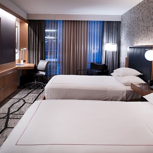 A modern hotel room with two beds, a TV, a desk, and large windows with curtains. The walls are decorated with textured wallpaper and artwork.