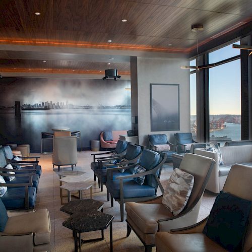 A modern, comfortable lounge area with a skyline mural, floor-to-ceiling windows offering waterfront views, and plush seating arranged in rows.