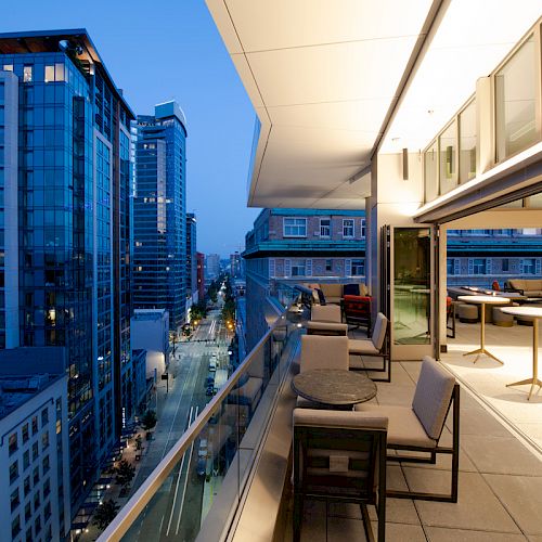 A modern cityscape with tall buildings is shown at dusk, featuring a rooftop patio with tables and chairs on the right side.