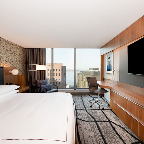 A modern hotel room with a large bed, a wall-mounted TV, a desk, and a view of city buildings through the window. The console is wooden.