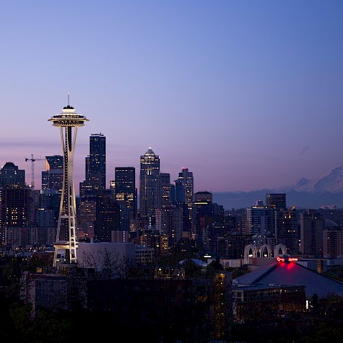 The image shows a nighttime city skyline featuring the Space Needle, high-rise buildings, and a distant mountain under a twilight sky.