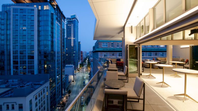 A modern balcony scene at dusk, with a cityscape view of tall buildings and a partially open interior space featuring tables and chairs.