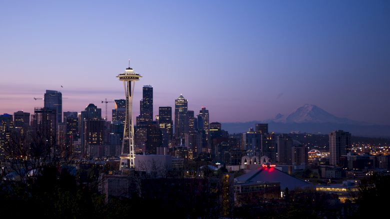 A twilight view of a city skyline featuring the Space Needle with a mountainous background and buildings illuminated.