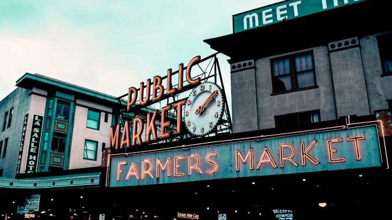 The image shows an iconic sign reading "Public Market" and "Farmers Market" with a clock, in a lively market area with buildings in the background.