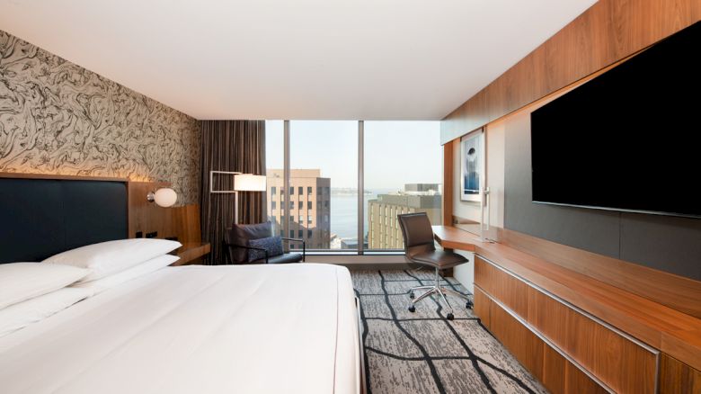 This image shows a modern hotel room with a large bed, a wall-mounted TV, a desk with a chair, large windows, and a view of buildings outside.