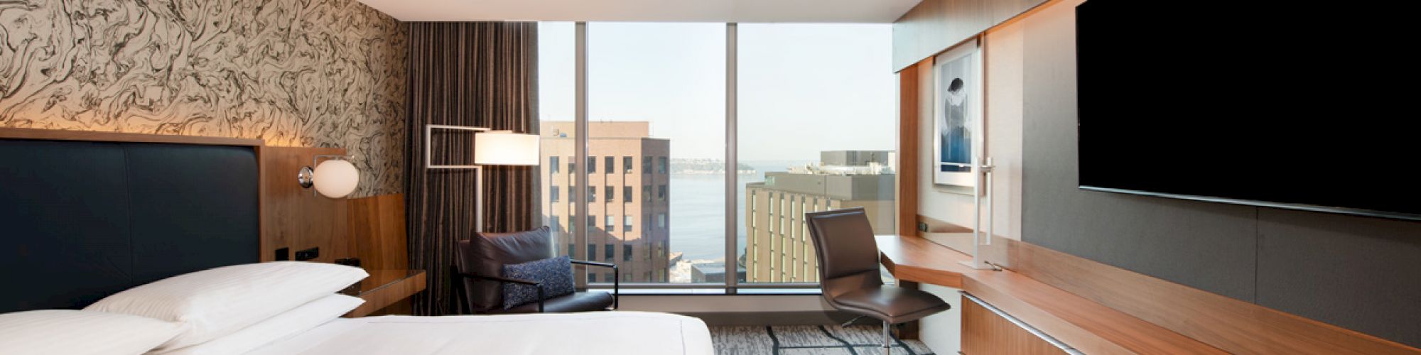 A modern hotel room with a king-sized bed, large TV, desk, chair, and a view of city buildings through floor-to-ceiling windows.