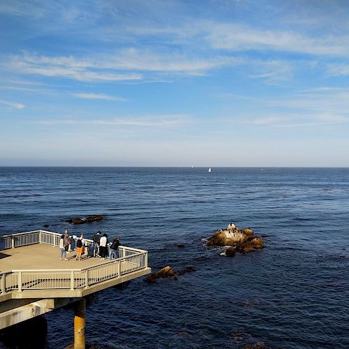 A group of people stand on a viewing platform overlooking a calm ocean with a small rocky outcrop and sailboats in the distance under a blue sky.