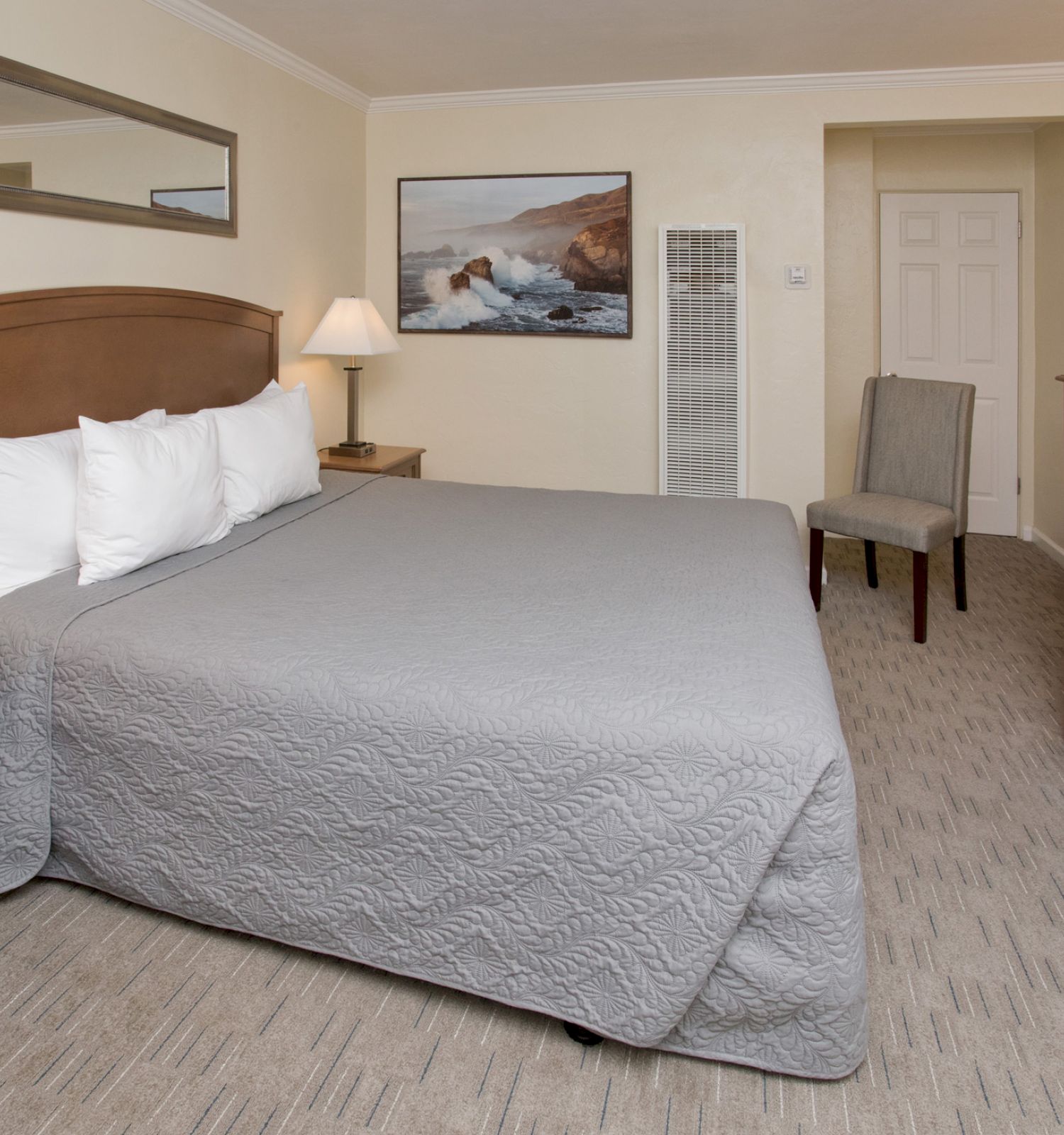 A cozy hotel room with a large bed, bedside tables with lamps, a chair, and a TV stand with a microwave and drawers. Wall art and a mirror adorn the space.