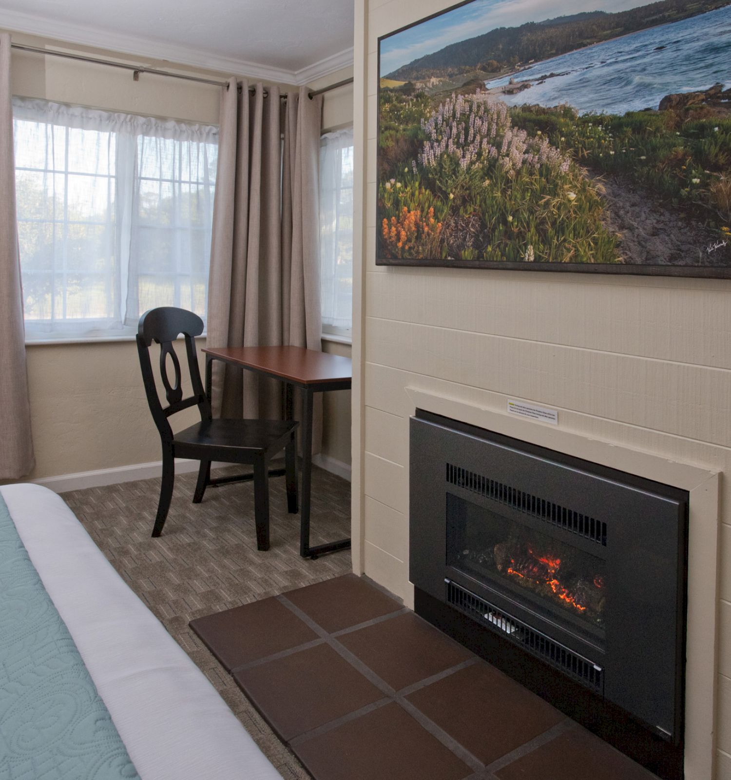 The image shows a cozy room with a bed, small table with a chair, a window, a wall-mounted fireplace and a painting above it.