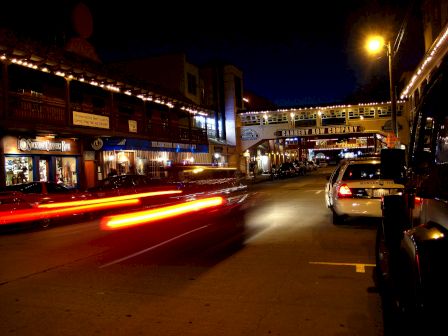 Night street scene with moving car light trails, storefronts illuminated, a bridge with lights above, and parked cars lining the street.