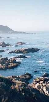 A coastal scene with rocky shores, calm blue waters, and a mountain in the background under a clear sky during the day.